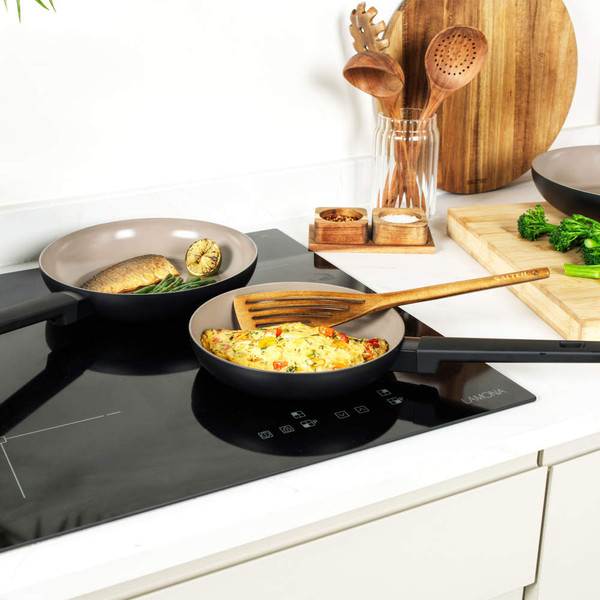 Two non-stick frying pans on an induction hob with cooked food, kitchen utensils in background