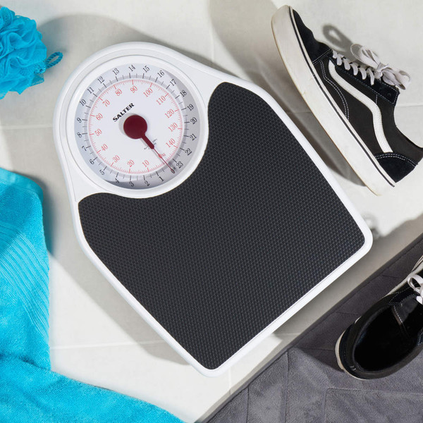 Mechanical bathroom scale with black platform, shown on tiled floor next to black trainers and blue towel