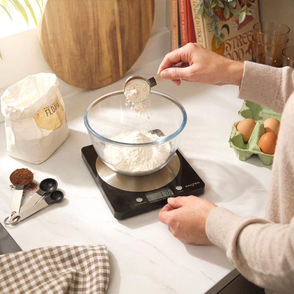 Digital kitchen scale weighing flour in glass bowl, baking ingredients nearby