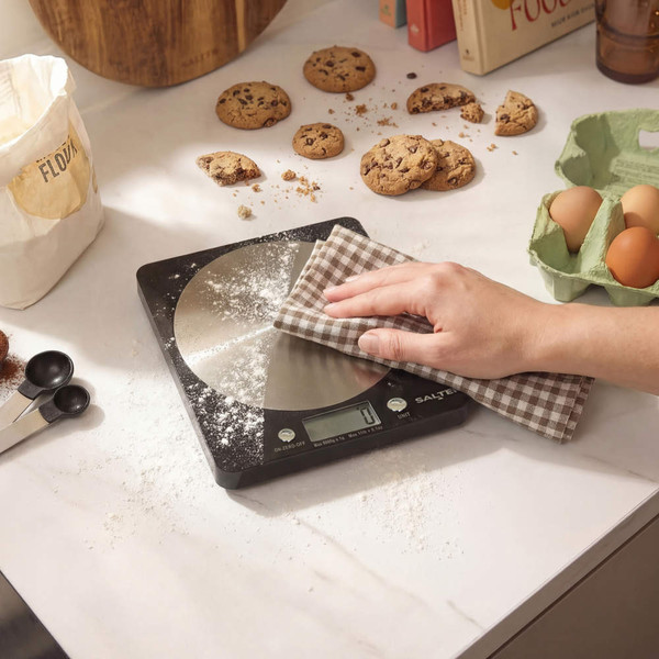 Digital kitchen scale being cleaned, surrounded by baking ingredients and cookies