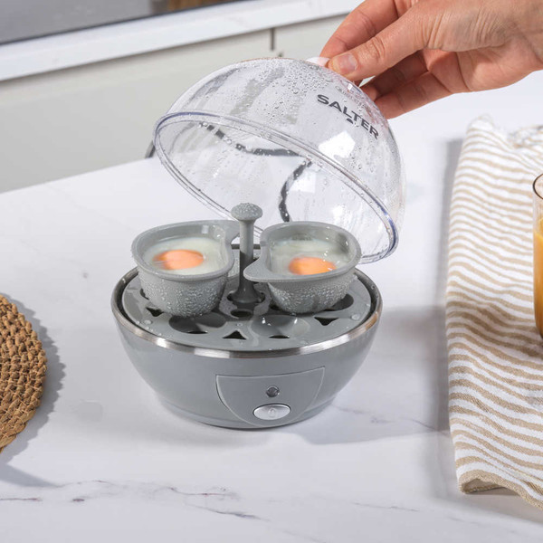 Electric egg cooker in grey with five brown eggs and a finger pressing the power button