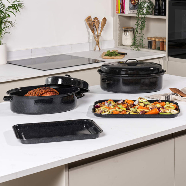 Black enamel roasting pan with lid, shown with cooked chicken and herbs inside on kitchen worktop.