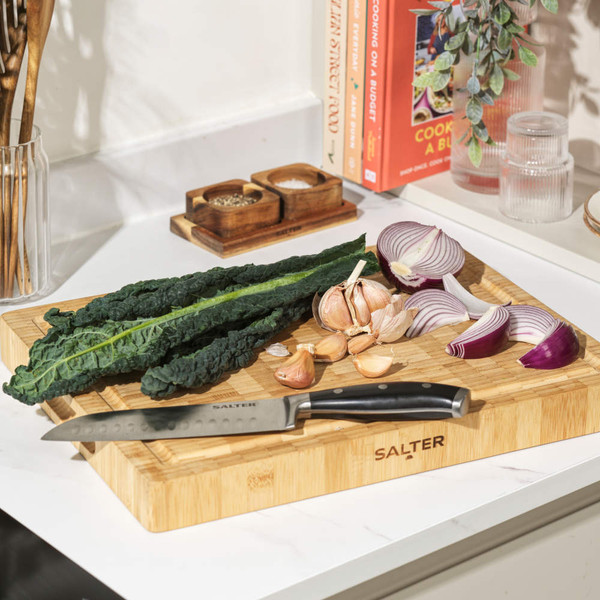 Chef’s knife slicing red onion on wooden Salter chopping board in kitchen.