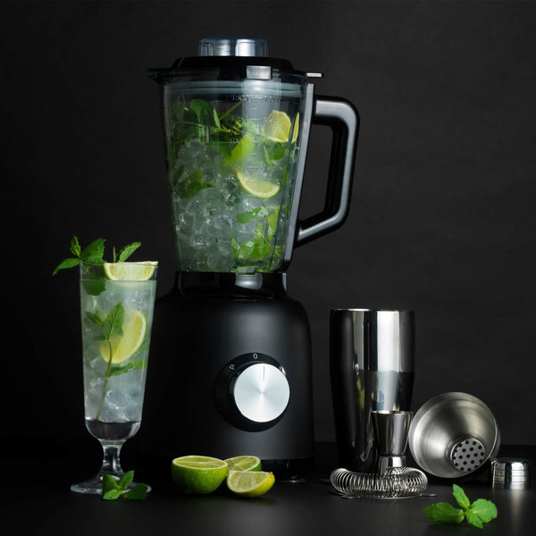 Black blender with jug of ice, lime and mint, cocktail glass and bar tools on black background