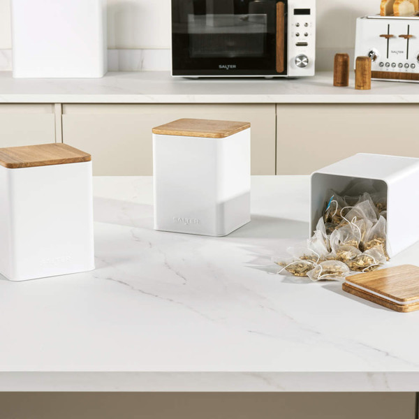 Set of three white kitchen storage canisters with wood lids on a counter, one holding teabags