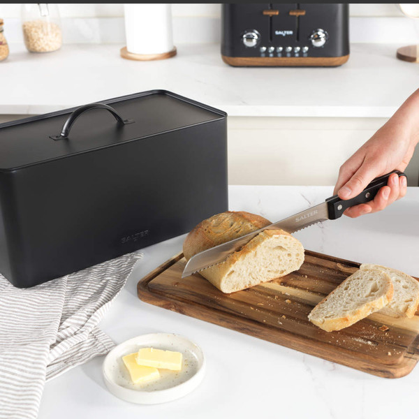 Black metal bread bin on kitchen counter beside bread and crumpets on wooden board