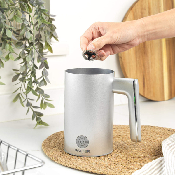 Silver electric milk frother with handle, shown in use on a kitchen counter
