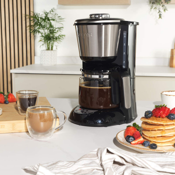 Black filter coffee maker with open lid showing ground coffee, next to a cup of cappuccino