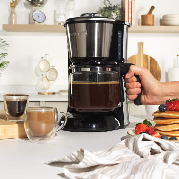 Black filter coffee maker with open lid showing ground coffee, next to a cup of cappuccino