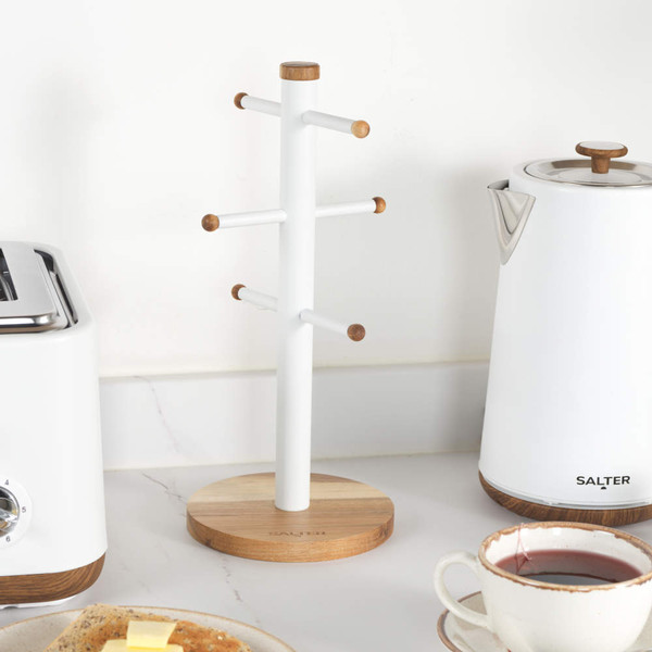 Hand placing cream ceramic mug with brown rim on white mug tree, next to white Salter kettle