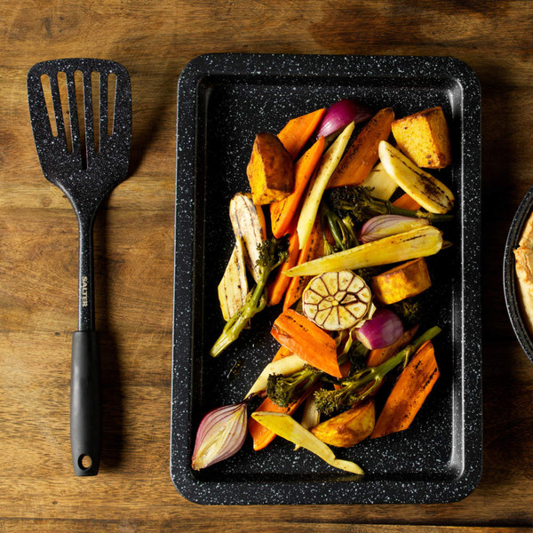 Roasting tray with assorted cooked vegetables beside black handled spatula on wooden surface