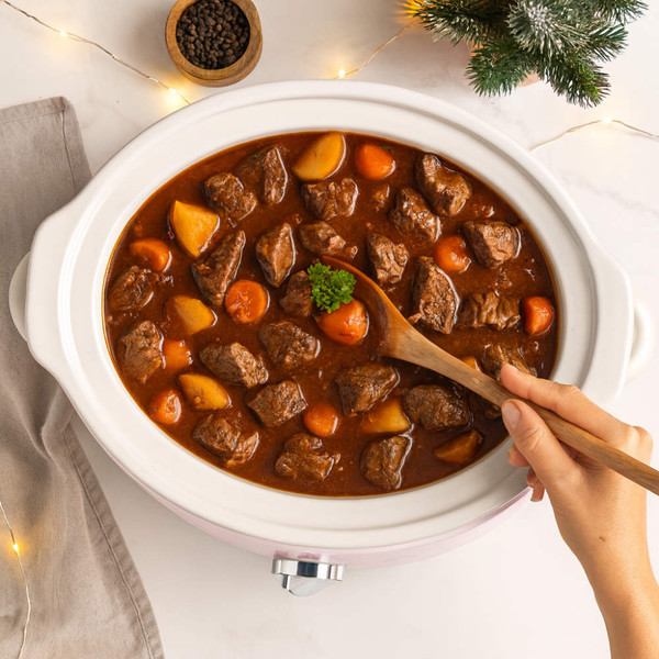 Slow cooker with beef stew, carrots and potatoes being stirred by hand