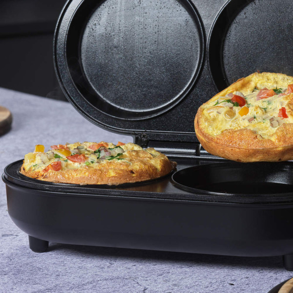 Black omelette maker with two compartments, one cooking vegetable omelette, on kitchen worktop