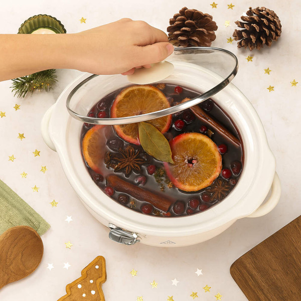 Cream slow cooker with festive mulled wine, orange slices, and spices on table.
