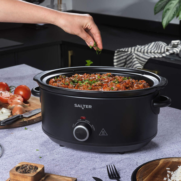 Salter black slow cooker filled with stew, hand adding herbs, on kitchen worktop