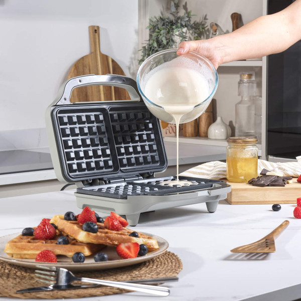 Electric waffle maker with batter being poured in, kitchen counter setup with waffles, berries and honey.
