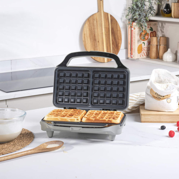Grey waffle maker in kitchen, batter being poured in, finished waffles with berries on plate