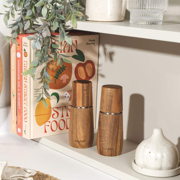 Walnut wood pepper mill upright on white worktop beside small bowl of black peppercorns