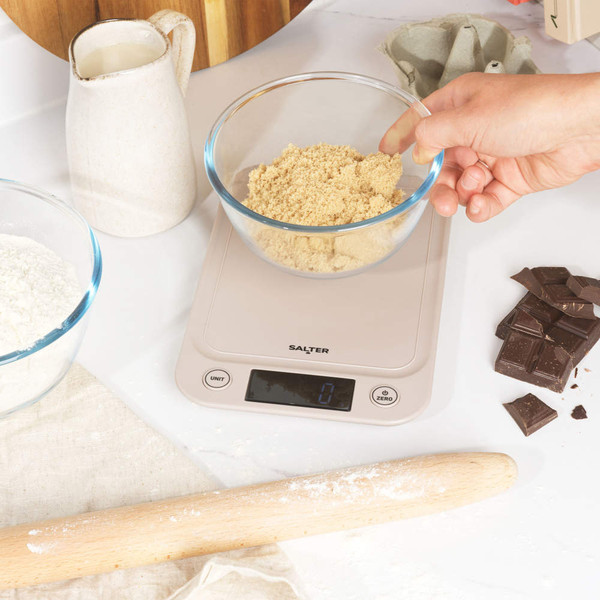 Digital kitchen scale with bowl of brown sugar being weighed, baking ingredients nearby
