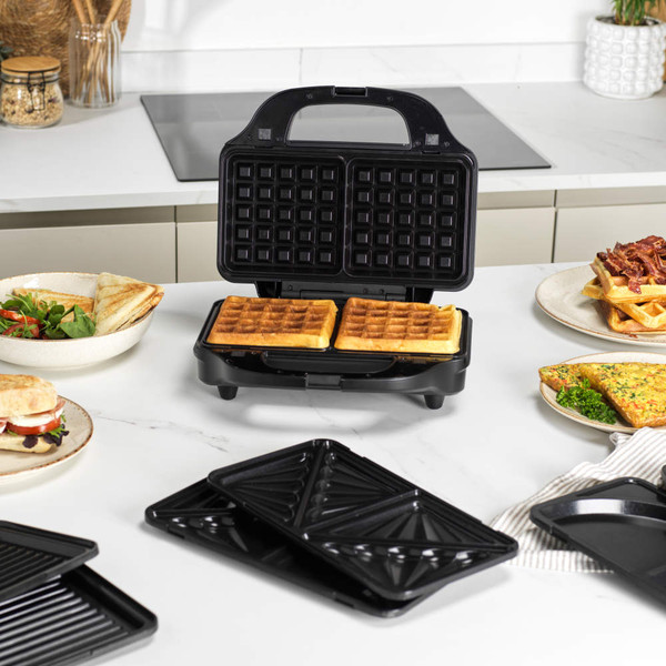 Black 3-in-1 sandwich maker open with waffle plates, shown on kitchen counter with waffles, toasties and sandwiches.
