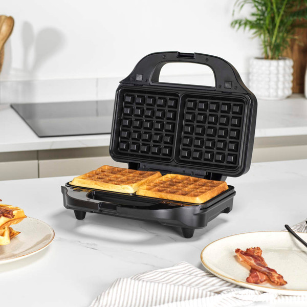 Black waffle maker open with two cooked waffles on kitchen worktop