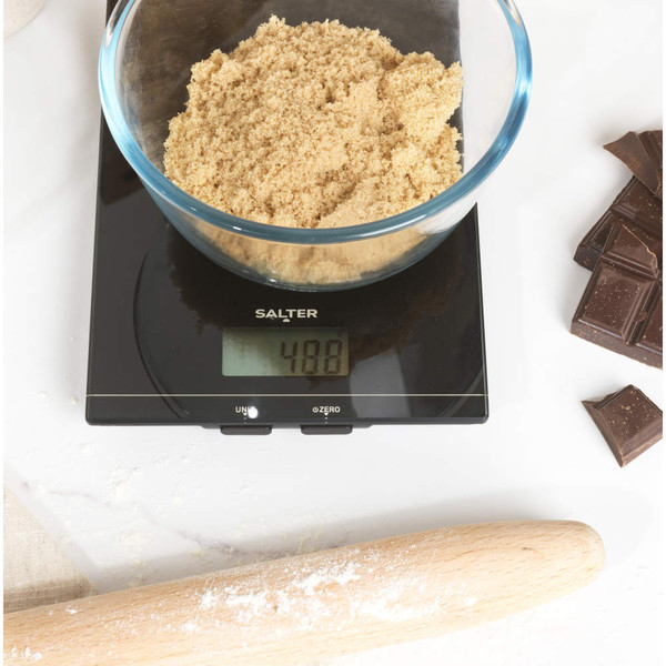 Glass bowl of brown sugar on black digital kitchen scale, chocolate pieces and rolling pin nearby