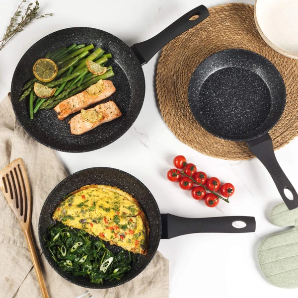 Set of two black speckled frying pans, one with omelette and greens, on kitchen counter
