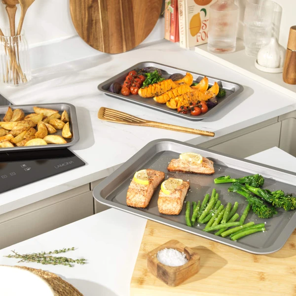 Non-stick oven trays with baked salmon, green veg, potato wedges and roasted tomatoes