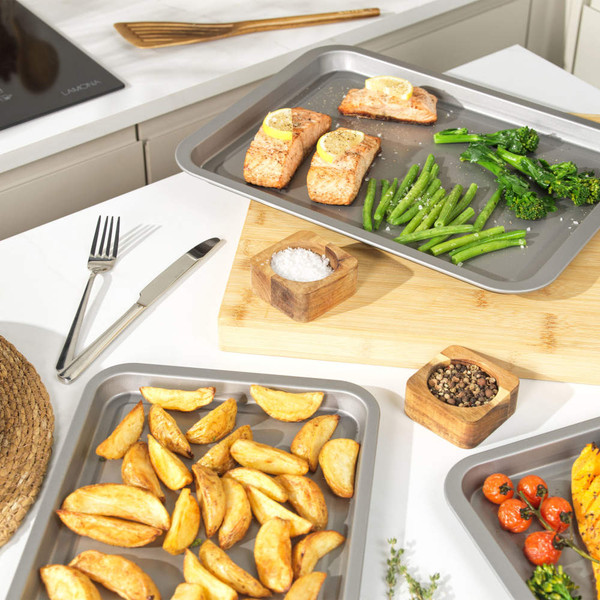 Non-stick oven trays with baked salmon, green veg, potato wedges and roasted tomatoes