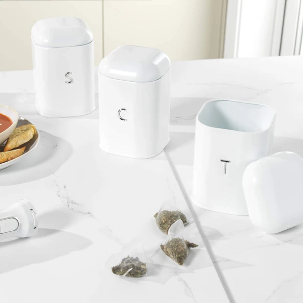Kitchen scene with hand taking tea from white canister, next to mug and fresh bread