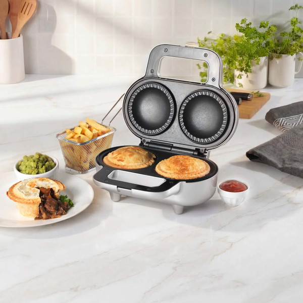 Silver pie maker with two baked pies, beside plate of pie, chips, peas and ketchup.