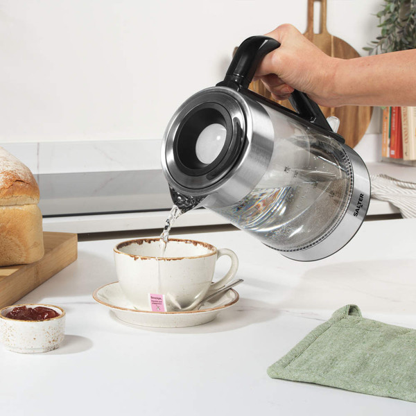 Glass electric kettle pouring hot water into teacup, kitchen worktop background