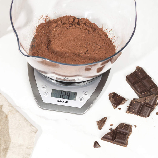 Digital kitchen scale weighing cocoa powder in clear bowl, with broken chocolate and eggs nearby.
