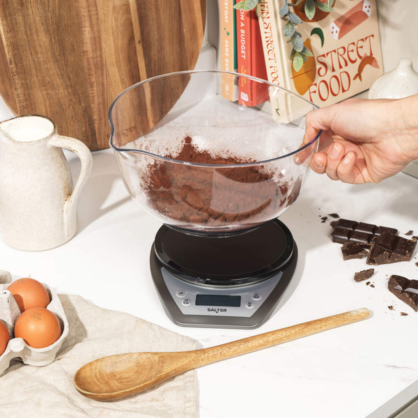 Digital kitchen scale with clear mixing bowl, cocoa powder, and hand pouring ingredients