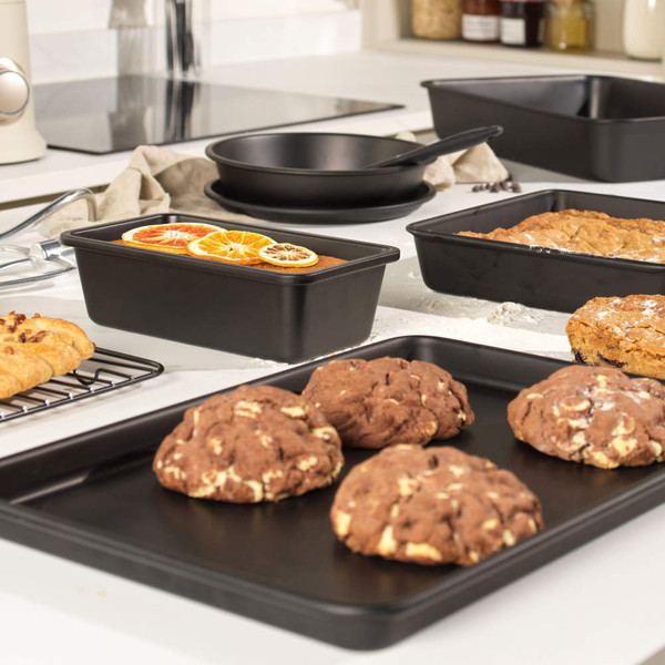 Black non-stick bakeware set displayed in kitchen with baked cookies, cake and croissants