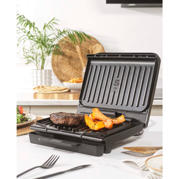 Electric grill with steak and vegetables cooking on kitchen counter, hand using spatula