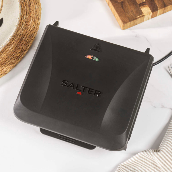 Black electric grill cooking steak and vegetables on kitchen counter, surrounded by plated food.