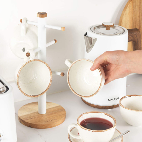 Hand placing cream ceramic mug with brown rim on white mug tree, next to white Salter kettle