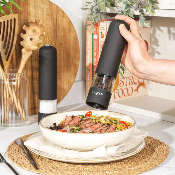 Black electric pepper grinder dispensing onto steak salad, with matching salt mill beside