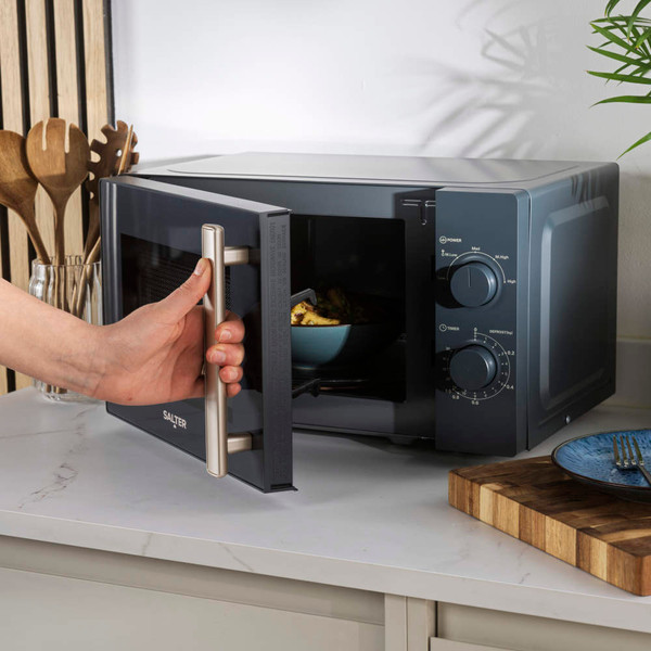 Grey microwave oven on kitchen counter, hand adjusting timer dial, plate and fork nearby