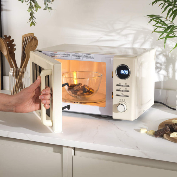 Cream microwave with digital timer set, hand turning dial, chocolate pieces on chopping board.