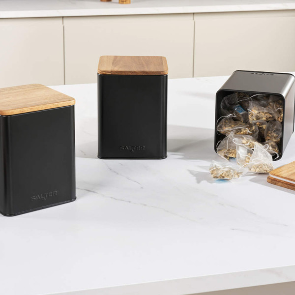 Set of black kitchen storage canisters with wooden lids, one open showing teabags.