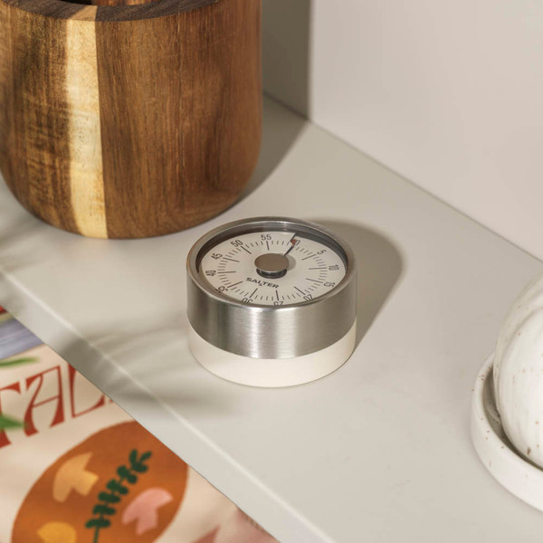 Cream and stainless steel mechanical kitchen timer with dial, shown on shelf.