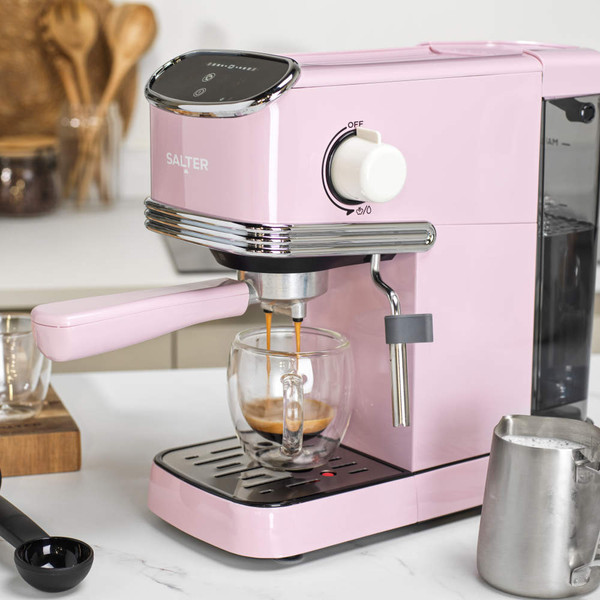 Pink espresso coffee machine in kitchen making coffee, shown with milk jug and scoop.