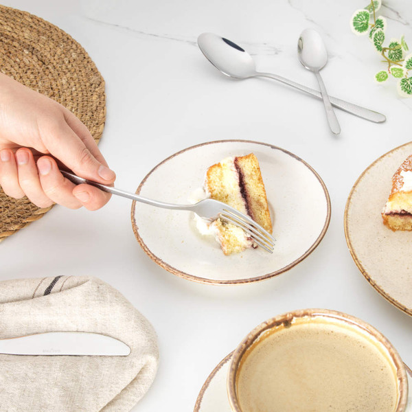 Hand holding fork with cake slice and cream on ceramic plate, coffee and cutlery nearby.