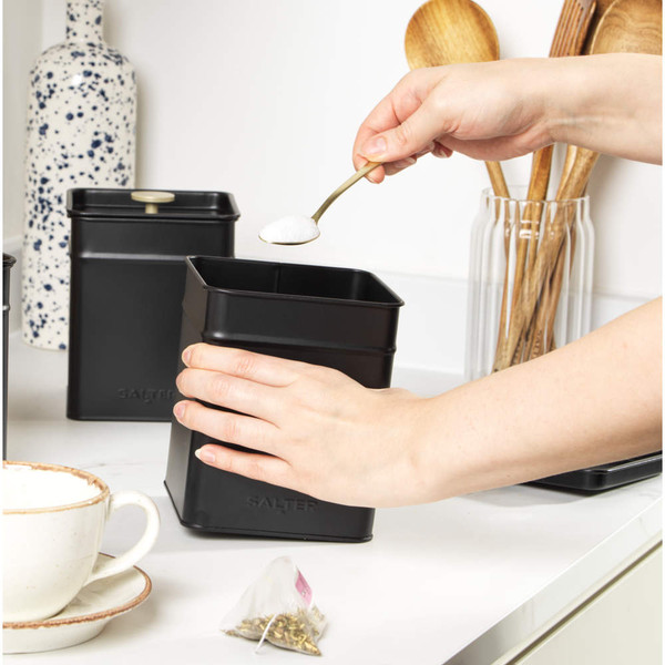 Hand adding sugar to black metal storage canister on kitchen counter