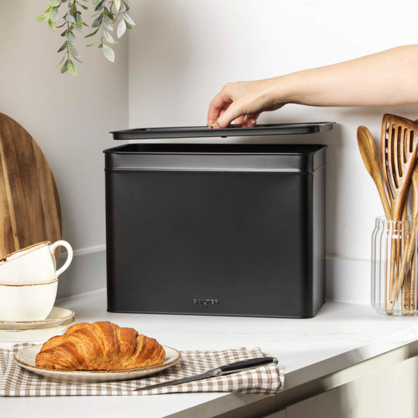 Black metal bread bin on kitchen counter, lid being lifted by hand