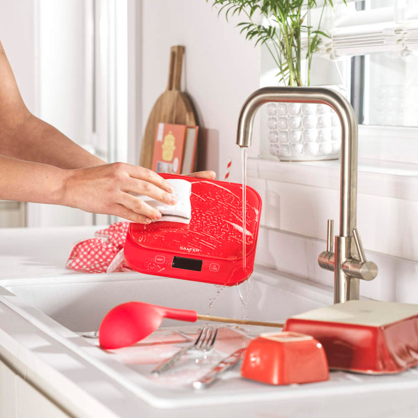 Red kitchen scale being wiped under running tap, showing waterproof cleaning feature