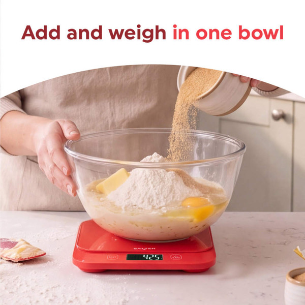 Red digital kitchen scale on countertop with strawberries, eggs, cake and glasses