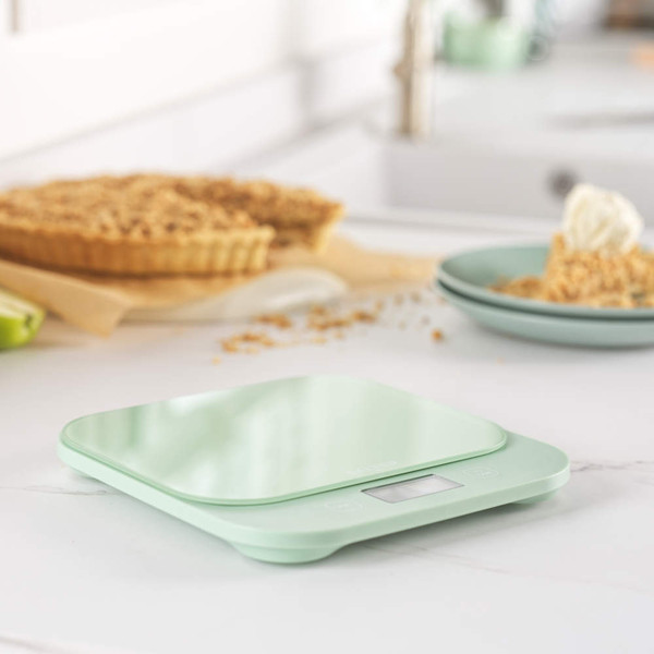 Digital kitchen scale in mint green with glass mixing bowl, apples, egg, butter and crumbs.